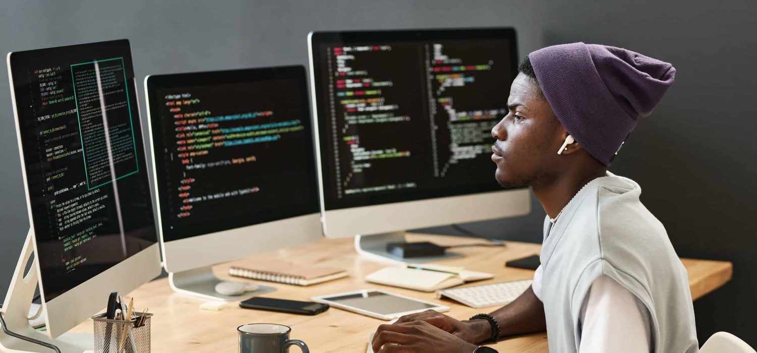 Young serious IT engineer in casualwear looking at coded data on computer screens while typing on keyboard by workplace